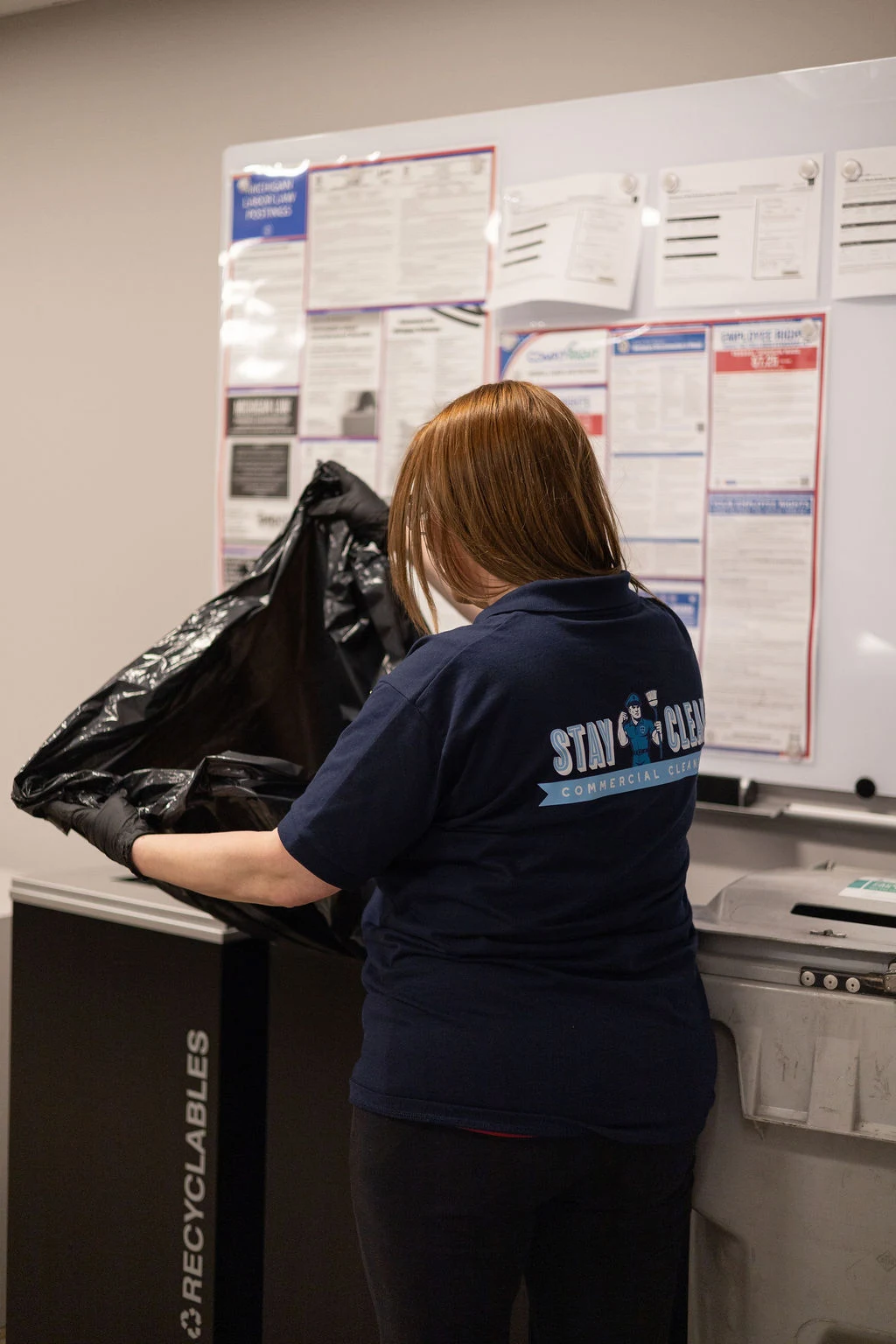 A Stay Clean team member handling waste and recycling beside a facility compliance board